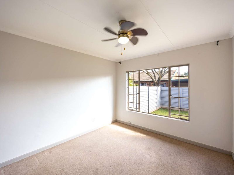 A bright, empty bedroom featuring neutral walls, carpeted flooring, and a ceiling fan, with a window overlooking the rear garden area.