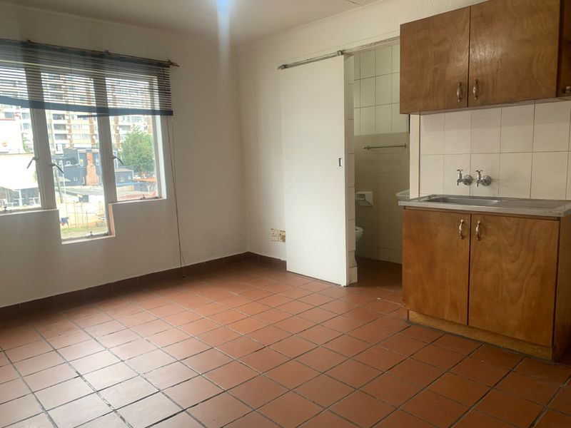 Open plan living area featuring tiled flooring and a compact kitchenette with wooden cabinetry, leading to an adjacent bathroom.