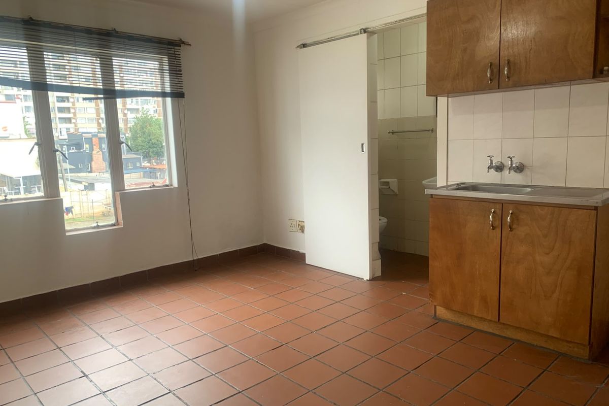 Open plan living area featuring tiled flooring and a compact kitchenette with wooden cabinetry, leading to an adjacent bathroom.