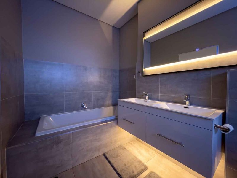 Modern bathroom featuring a sleek double vanity with integrated basins, a large backlit mirror, and a built-in bathtub with ambient under-cabinet lighting.