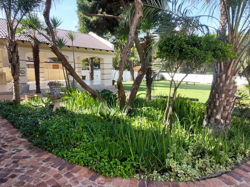 A well-maintained rear garden featuring a brick-paved pathway, mature landscaping, and a spacious covered patio area with a built-in braai.