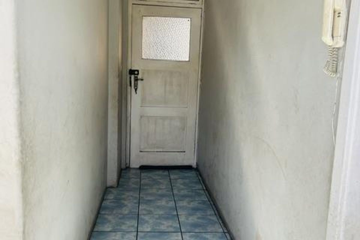 A narrow hallway featuring tiled flooring and an intercom system, leading to a panelled entrance door.