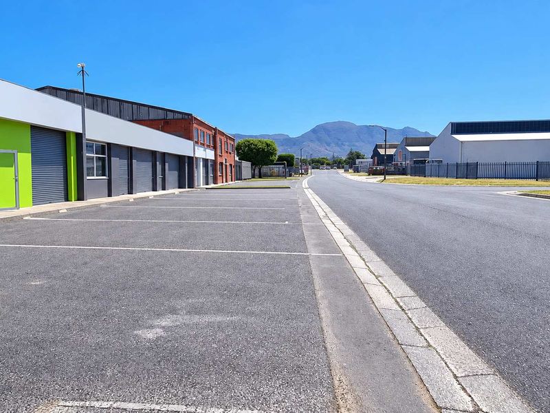 A clear street-level view of an industrial park featuring paved parking bays, modern building facades with roller shutter doors, and perimeter security fencing.