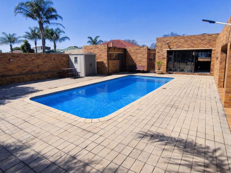 A well-maintained outdoor pool area featuring a rectangular swimming pool, extensive paved decking, and a brick boundary wall with an adjacent covered patio.