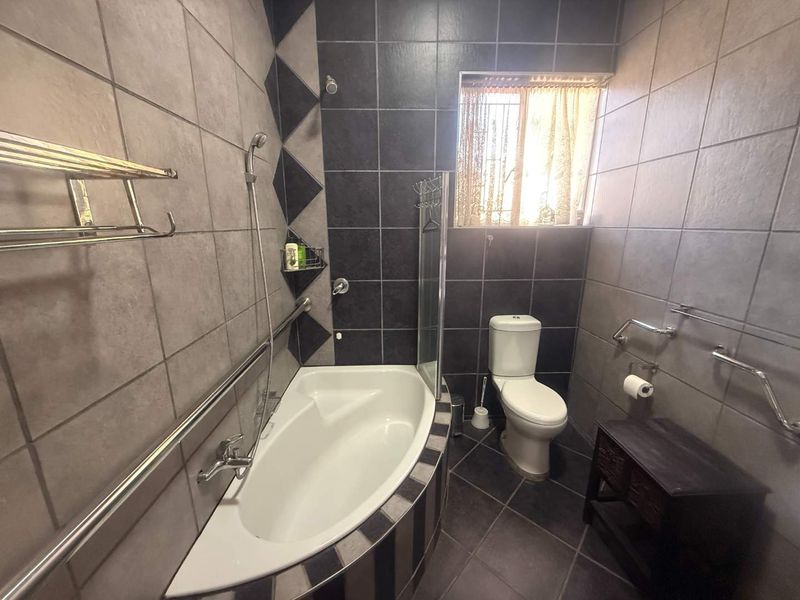 A functional bathroom featuring a corner bathtub with handheld shower, toilet, and safety grab rails, finished with dark and grey wall and floor tiling.