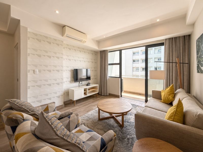 A well-maintained living room featuring tiled flooring, recessed lighting, a wall-mounted air conditioner, and direct access to a private balcony through sliding glass doors.