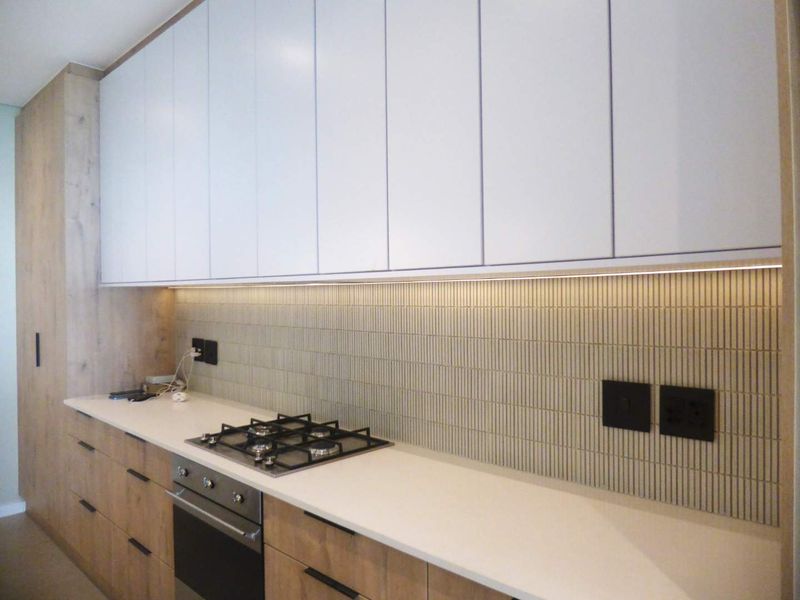 Modern kitchen featuring sleek white upper cabinetry, wood-grain lower cabinets, stone countertops, a gas hob, and a stylish vertical tile backsplash.