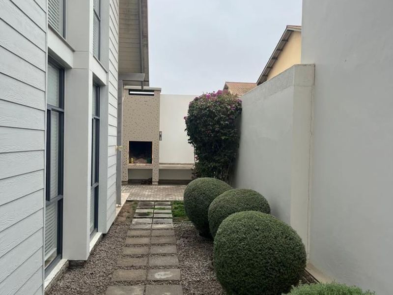 A side pathway leading to a rear outdoor entertainment area featuring a built-in braai, paved walkway, and landscaped garden beds.
