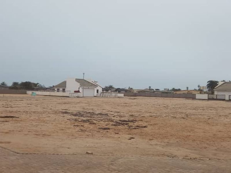 A large, undeveloped plot of land featuring sandy terrain, bordered by boundary walls and adjacent residential properties.