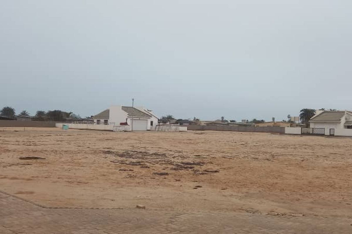 A large, undeveloped plot of land featuring sandy terrain, bordered by boundary walls and adjacent residential properties.