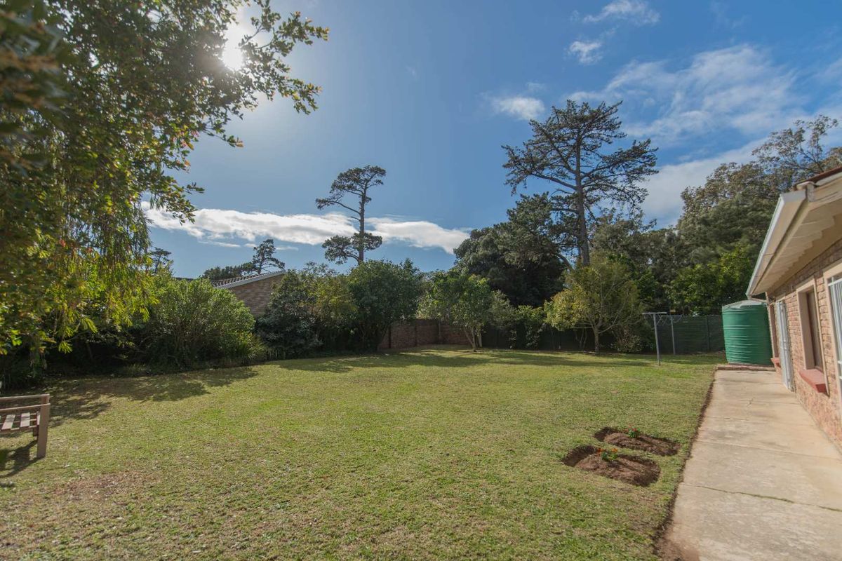 A spacious rear garden featuring a well-maintained lawn, mature trees, perimeter fencing, and a concrete pathway along the side of the house.