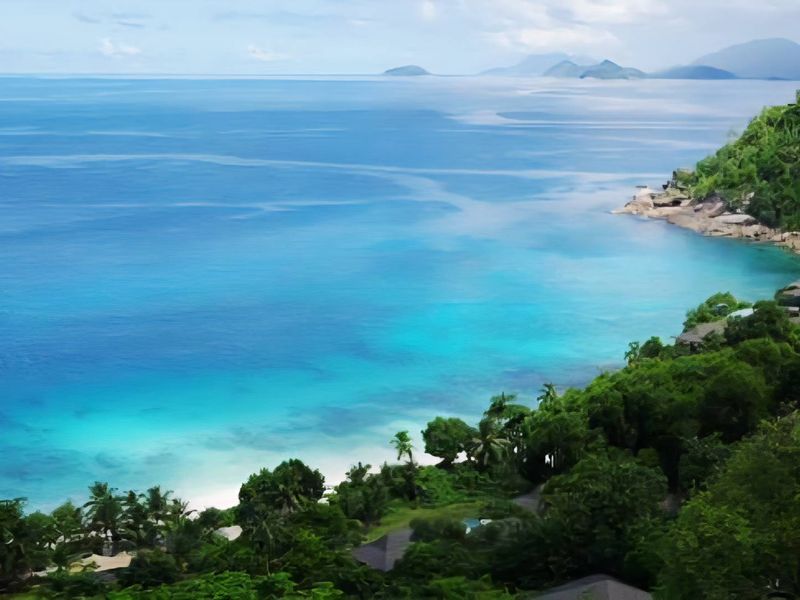 A stunning elevated aerial view of a coastal landscape featuring lush tropical vegetation, a pristine beach, and expansive turquoise ocean waters.