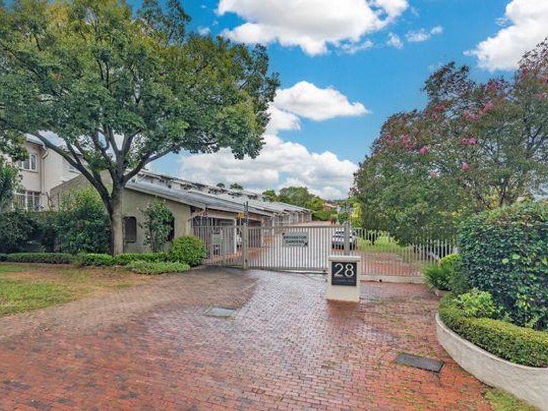 Front entrance view of the townhouse complex featuring a brick paved driveway, secure metal gate, and established garden landscaping.