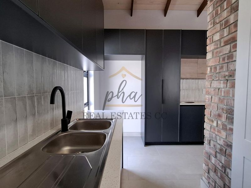 Modern kitchen featuring sleek dark cabinetry, a double bowl sink with black fixtures, tiled backsplash, and an exposed brick feature wall.