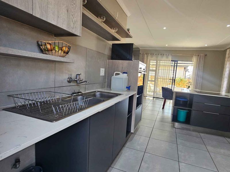 Modern kitchen featuring sleek dark cabinetry, stone countertops, built-in oven, and tiled flooring, with an open-plan layout leading to the outdoor area.