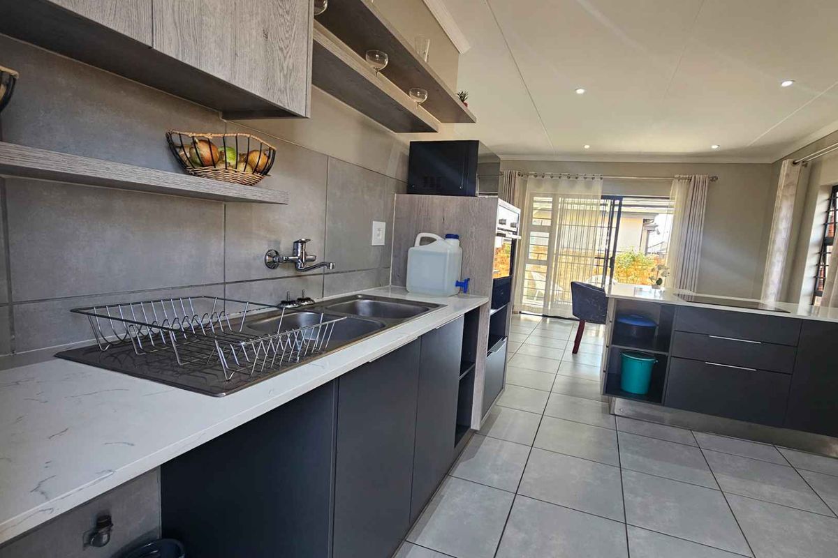 Modern kitchen featuring sleek dark cabinetry, stone countertops, built-in oven, and tiled flooring, with an open-plan layout leading to the outdoor area.