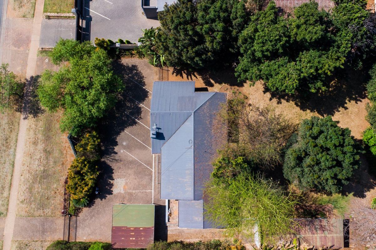 Aerial view of the property showing the main building with a metal roof, paved parking area, and surrounding mature garden landscaping.
