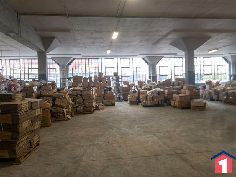 Spacious industrial warehouse area featuring concrete flooring, structural columns, and large steel-framed windows providing ample natural light.