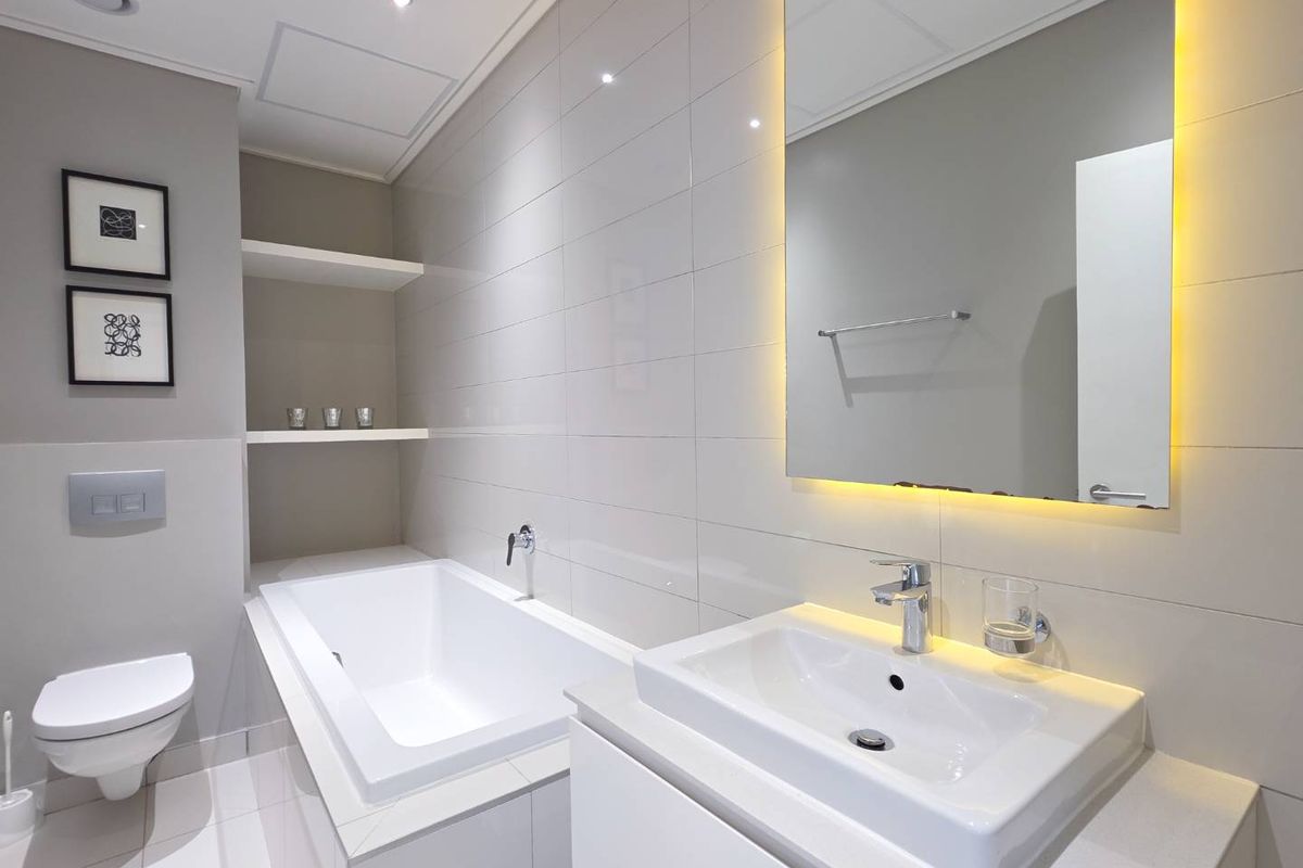 Modern bathroom featuring a wall-mounted toilet, built-in bathtub, vanity basin with backlit mirror, and recessed shelving with contemporary tiled finishes.