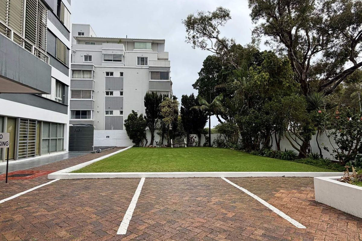 A well-maintained communal outdoor area featuring a paved parking space, a neat lawn, and established garden landscaping adjacent to the apartment building.