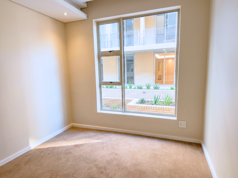 A bright, empty bedroom featuring neutral carpeted flooring, recessed ceiling lighting, and a large window overlooking the complex courtyard.