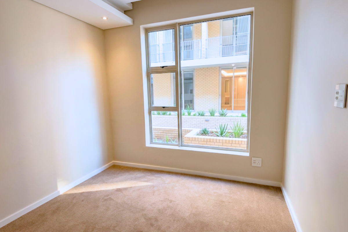 A bright, empty bedroom featuring neutral carpeted flooring, recessed ceiling lighting, and a large window overlooking the complex courtyard.