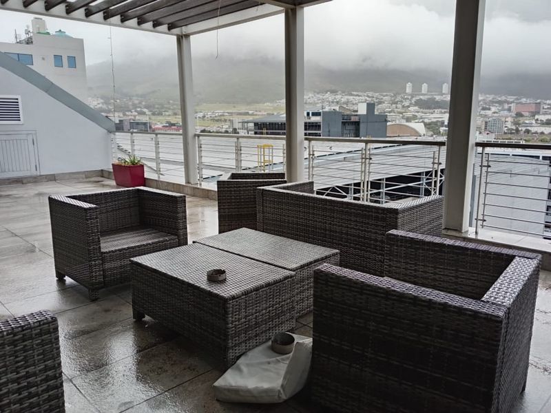 A covered outdoor balcony featuring tiled flooring, a pergola structure, and metal railings, offering expansive views of the surrounding urban landscape.
