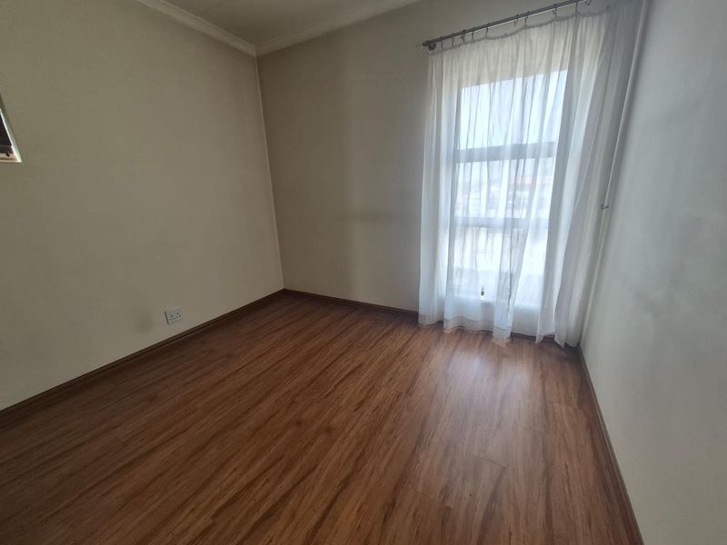 A bright, empty bedroom featuring warm-toned laminate flooring, neutral walls, and a large window with a fitted curtain rod.