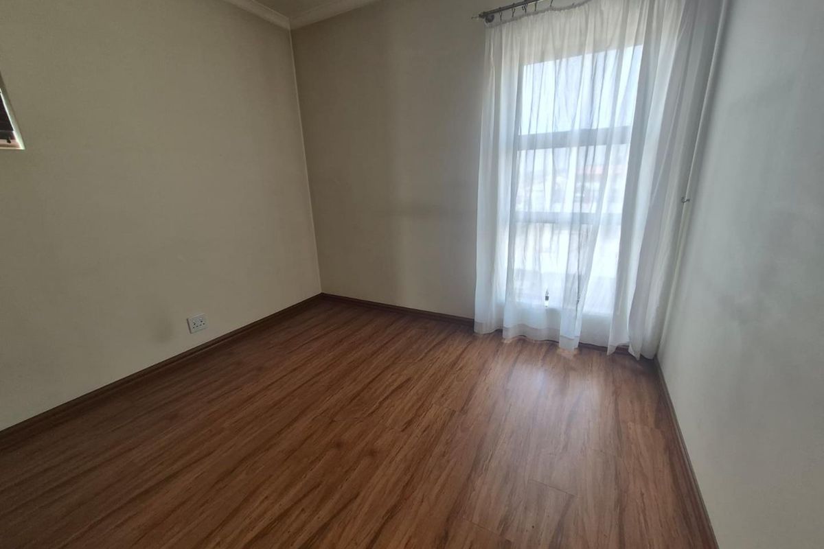 A bright, empty bedroom featuring warm-toned laminate flooring, neutral walls, and a large window with a fitted curtain rod.