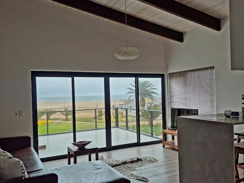 Open-plan living area featuring exposed timber ceiling beams, concrete kitchen island, and large sliding glass doors opening to a scenic beach view.
