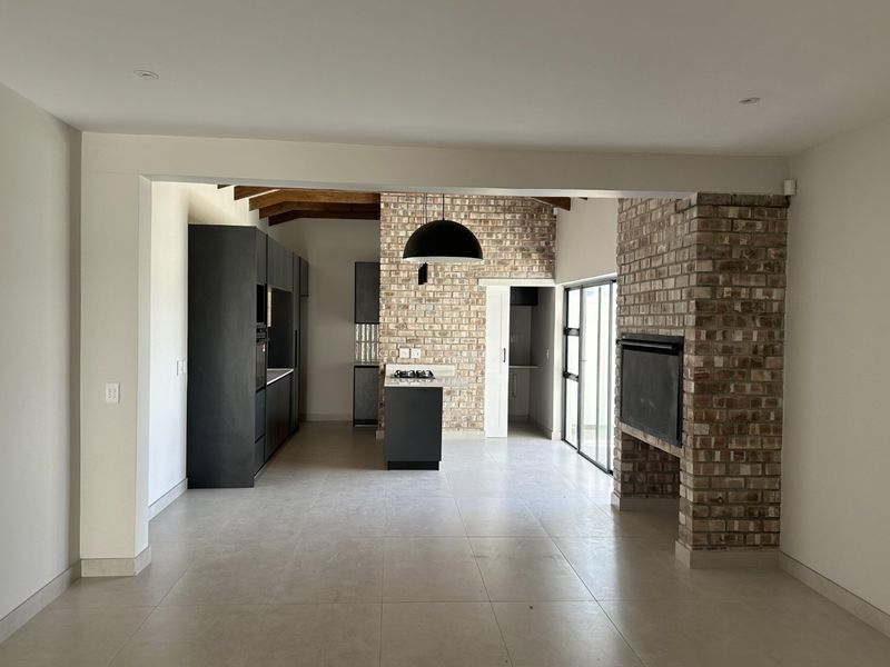Spacious open-plan living and kitchen area featuring exposed brickwork, timber ceiling beams, a built-in fireplace, and modern dark cabinetry with a central island.