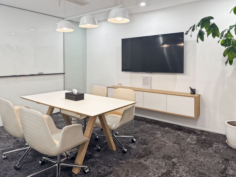 A modern, well-lit office meeting room featuring a glass whiteboard, wall-mounted storage cabinetry, and professional carpeted flooring.
