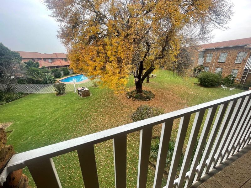 Elevated balcony view overlooking a communal garden area featuring a swimming pool, mature trees, and a brick braai facility.