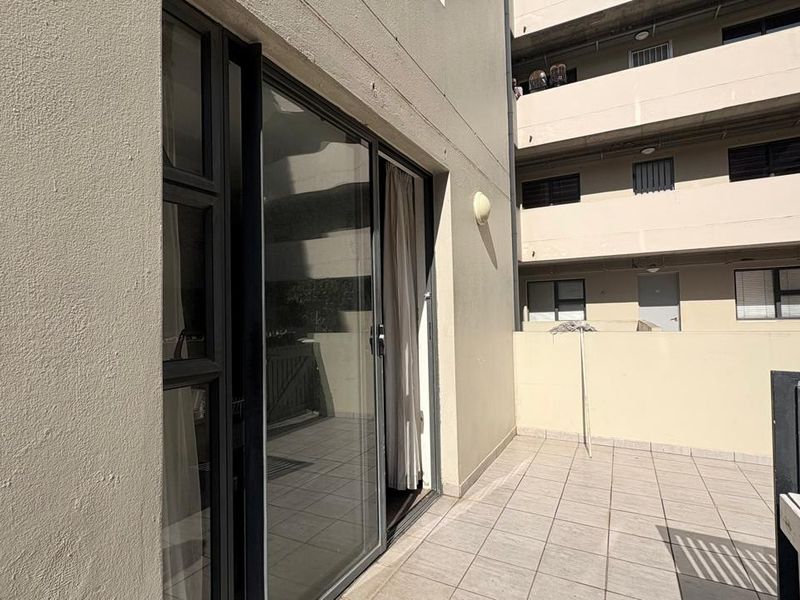 A tiled balcony area featuring a sliding glass door entrance and a simple balustrade, overlooking the apartment complex courtyard.