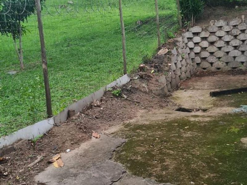 Rear outdoor area featuring a concrete paved section, retaining wall, and perimeter fencing with razor wire, set against a grassy slope.
