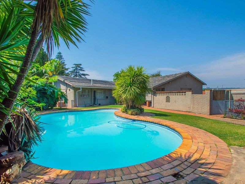 A well-maintained outdoor pool area featuring a curved brick-edged swimming pool, established garden, and brick boundary walls.