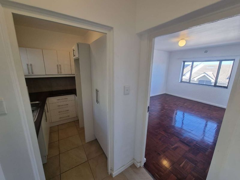 An open-plan view from a hallway into a kitchen with white cabinetry and a bright, empty living area featuring polished parquet flooring.