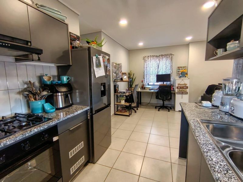 Open-plan kitchen featuring modern grey cabinetry, granite countertops, and integrated appliances, flowing into a functional workspace area with tiled flooring throughout.