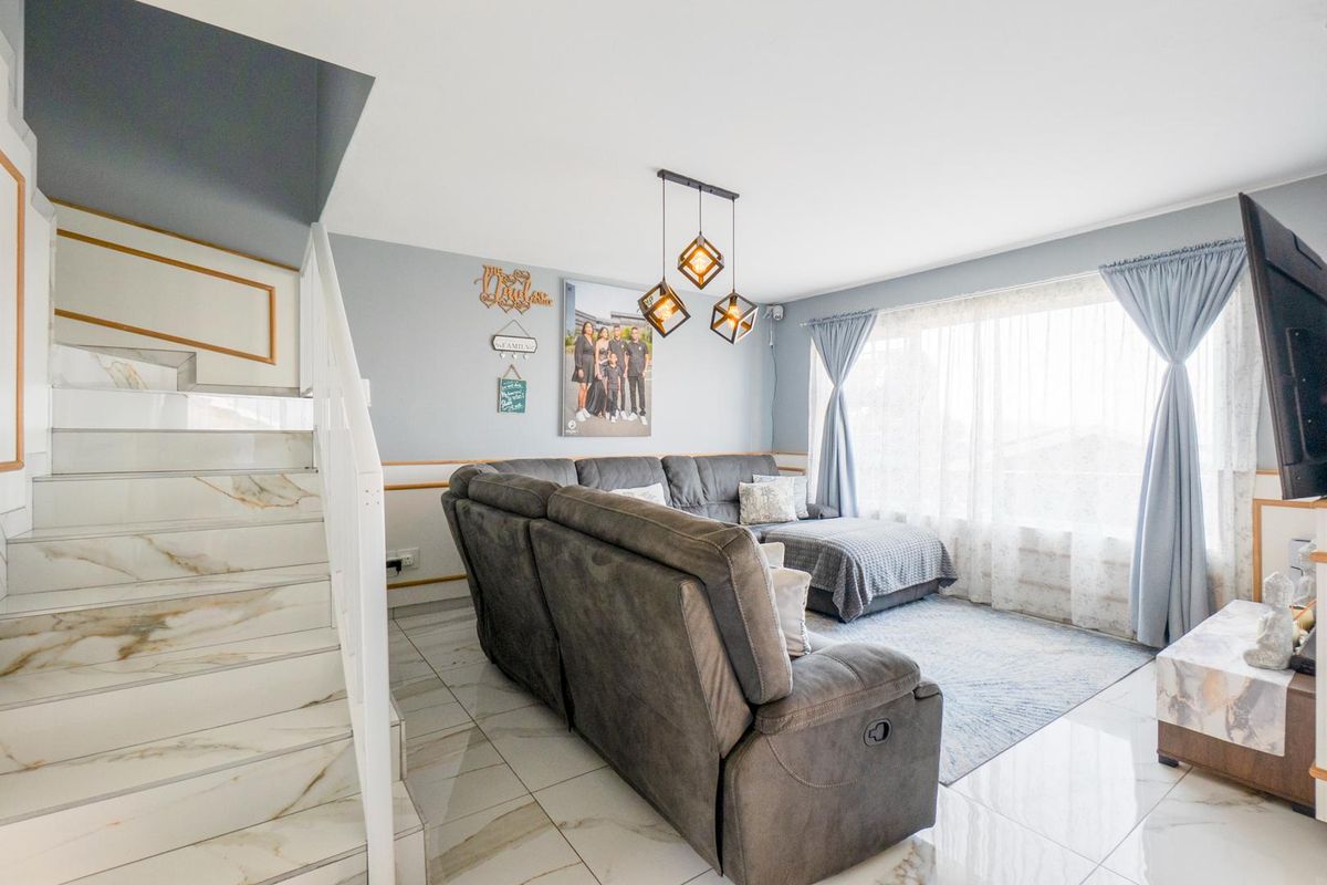 A bright living room featuring polished floor tiles, a marble-look staircase with a white balustrade, and large windows allowing ample natural light.