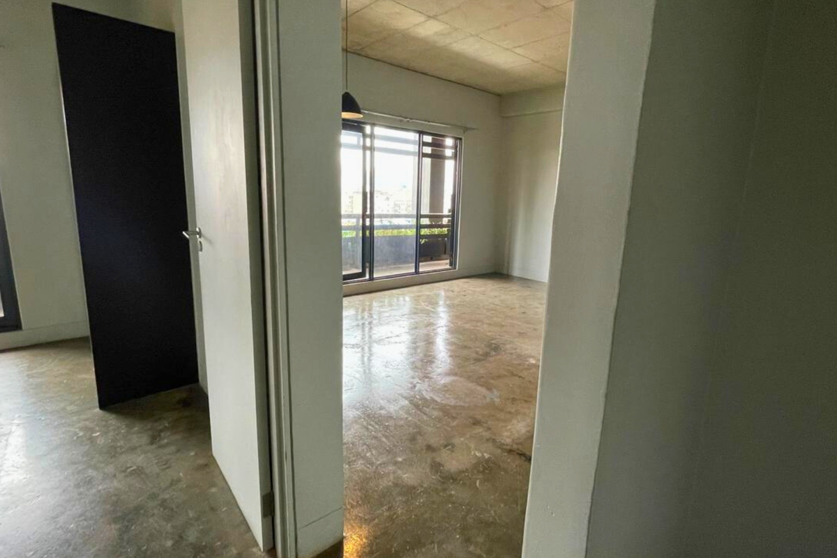 An empty, industrial-style apartment interior featuring raw concrete ceilings and polished concrete floors with large sliding glass doors leading to a balcony.