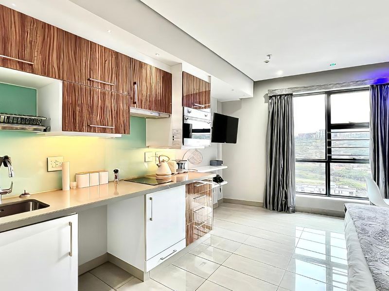 Modern open-plan kitchen featuring wood-grain cabinetry, integrated appliances, and sleek tiled flooring, seamlessly connected to a bright living area with large windows.
