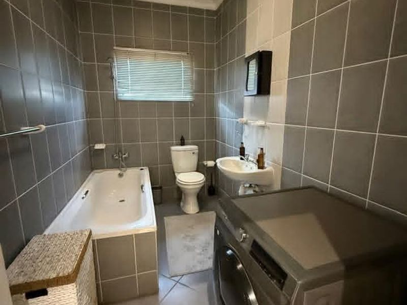 A functional bathroom featuring a built-in bathtub, wall-mounted basin, and toilet, finished with neutral grey and white wall tiling.