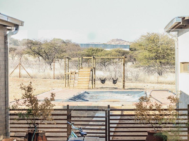 Outdoor pool area featuring a covered swimming pool, wooden jungle gym, and metal perimeter fencing set against a natural landscape.