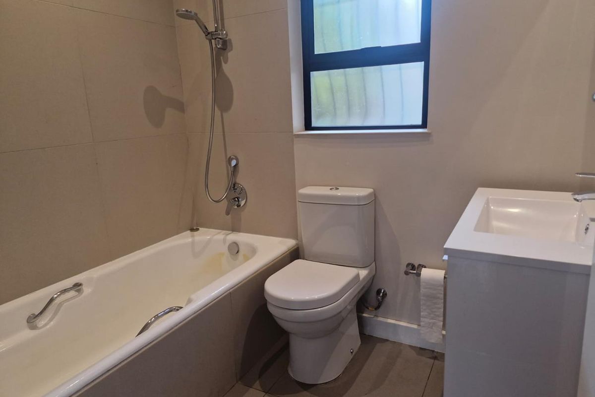A functional bathroom featuring a bathtub with handheld shower, a toilet, and a vanity basin with neutral wall and floor tiling.
