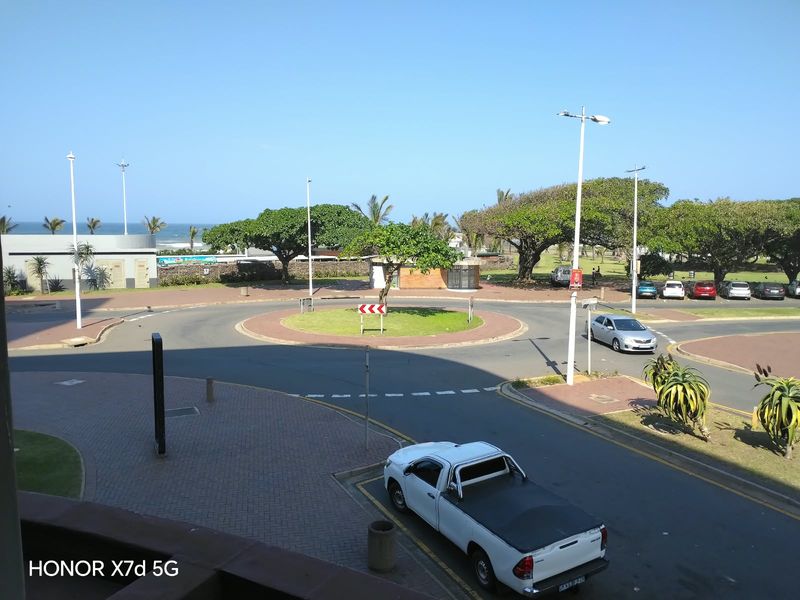 An elevated view overlooking a coastal roundabout, street infrastructure, and surrounding landscaped areas with a glimpse of the ocean in the distance.