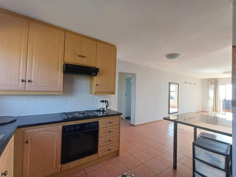 Open-plan kitchen and living area featuring wooden cabinetry, integrated appliances, and tiled flooring, leading to a bright living space with balcony access.