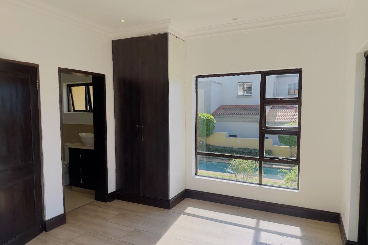 A bright bedroom featuring built-in dark wood cabinetry, tiled flooring, and an en-suite bathroom, with large windows overlooking the pool area.