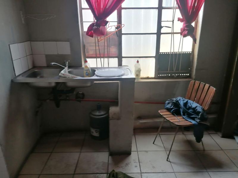 A basic kitchen area featuring a stainless steel sink, tiled splashback, and tiled flooring, requiring significant cosmetic updates and maintenance.