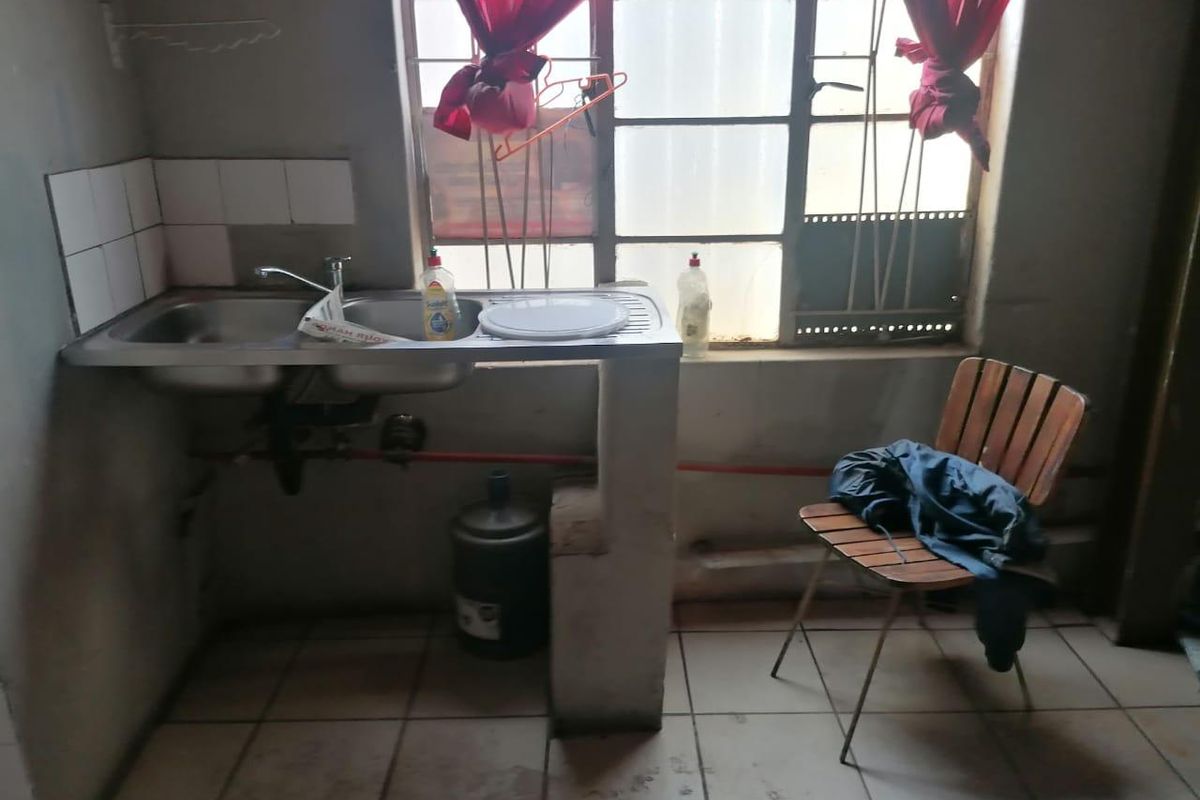 A basic kitchen area featuring a stainless steel sink, tiled splashback, and tiled flooring, requiring significant cosmetic updates and maintenance.