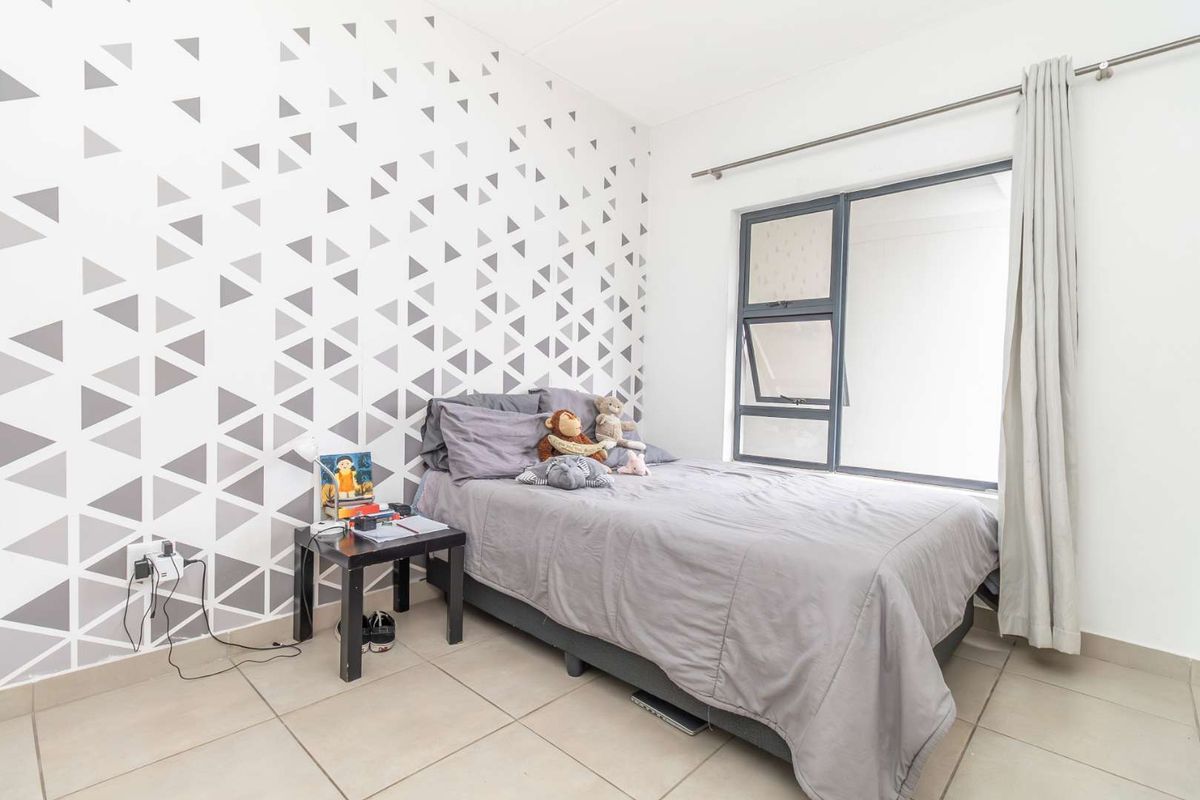 A bright bedroom featuring tiled flooring, a modern geometric patterned accent wall, and a large window with fitted curtain rails.